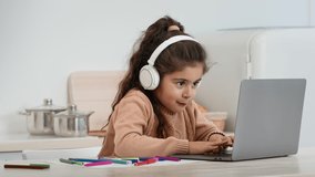 Little Arabic Girl Using Laptop Playing Games Online Wearing Headphones Sitting In Kitchen At Home. Kid And Gadget Concept. E-Learning, Modern Technology Concept. Side View Shot, Zoom In