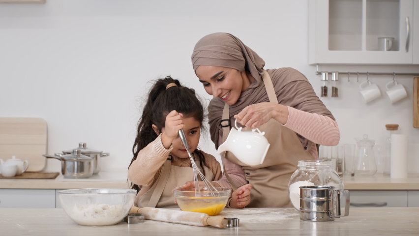 Muslim Family Baking In Kitchen. Happy Middle-Eastern Mom In Hijab And Her Little Daughter Making Dough Adding Eggs And Milk And Mixing Ingredients In Bowl Sitting In Kitchen At Home. Zoom In Royalty-Free Stock Footage #1073810219