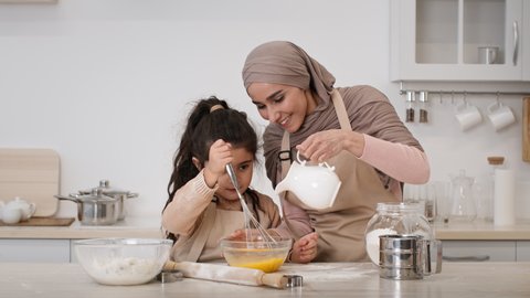 Muslim Family Baking In Kitchen. Happy Middle-Eastern Mom In Hijab And Her Little Daughter Making Dough Adding Eggs And Milk And Mixing Ingredients In Bowl Sitting In Kitchen At Home. Zoom In