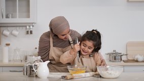 Cheerful Muslim Mother In Hijab And Her Daughter Baking Together Making Dough Mixing Eggs In Bowl In Modern Kitchen Indoors. Middle-Eastern Family Of Two Cooking Cookies On Weekend. Zoom In