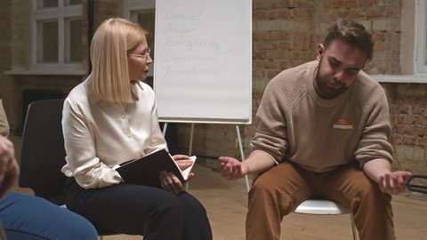 Medium shot of young slovenly man with beard and messy hair talking about his mental or addiction problem to female psychologist making notes in notebook during group therapy session