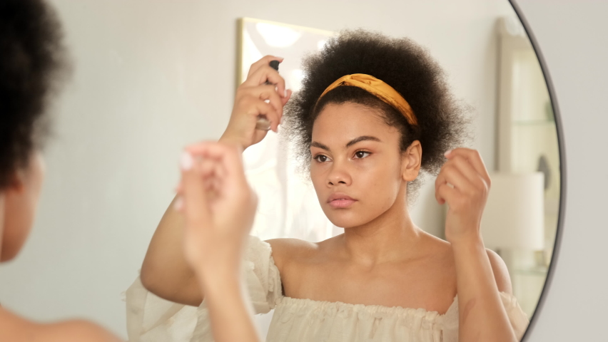 African american woman applies hair spray care balm styling cosmetics on damaged dry ends, makes hairstyle in front of the mirror at home. Royalty-Free Stock Footage #1073823329
