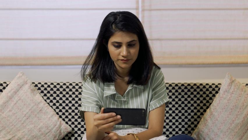 Beautiful Indian girl holding smart phone watching funny videos,movie or social media content while sitting in home office.Pretty woman looking at phone screen and watching something funny indoor. Royalty-Free Stock Footage #1073823935