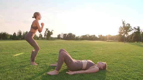 two women doing sports exercises on green grass in the park. Squats and abdominal exercises. young people lead an active sports lifestyle. girls have yoga classes. maintaining a healthy body and mind.