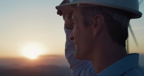 Cinematic close up of young engineer putting security helmet on satisfied with his job on background of wind mills with at sunset.Concept: renewable energy,technology,electricity,service,green, future