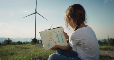 Cinematic shot of happy little girl is drawing wind turbines and enjoying view on windmill field with sun shining. Concept of renewable energy, love for nature, family, electricity, green, future.
