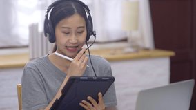 Young Asian female agent wear headset chatting and consultant as call center sit in living room using digital tablet and laptop while working at home advice as customer service