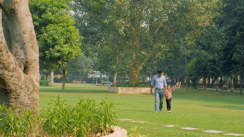 Urban father wearing shirt and jeans enjoying a leisure walk with his daughter. Long shot of a beautiful park where a dad having a fun time with his little girl - togetherness and bonding concept
