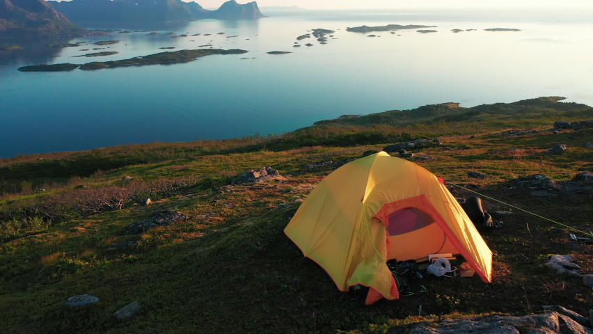 Aerial view over a tent on a mountain ledge, Bergsoyan archipelago background, midnight sun, in Senja, Norway, - reverse, tilt up drone shot Royalty-Free Stock Footage #1073832938
