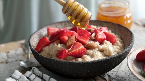 Pouring honey on oatmeal porridge served with fresh strawberries and almonds. Healthy breakfast food, clean eating, dieting concept