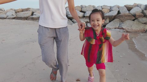 Adorable asian 5 years old girl and her mother are playing, running and chasing camera with smile and laughing to express her emotion on the beach near the sea and strong wind shows cheerful feeling.