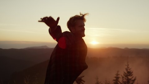 Excited man dancing in mountains at sunset. Happy guy gesturing hands during dance. Smiling male hiker jumping in air at summer weekend. Positive man jumping in air 