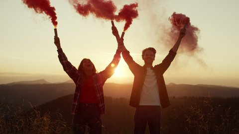 Happy woman and man with smoke bombs standing in meadow. Excited couple waving active smoke grenades in mountains at sunset. Positive guy and girl having fun. Hipsters running through colorful smoke