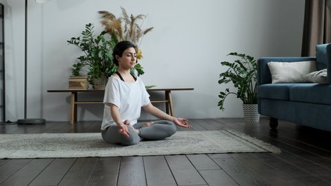 Pretty young Indian woman is practicing yoga at home relaxing in simple body position sitting on floor with beautiful furniture around. The girl is wearing a white T-shirt and black leggings. Healthy 