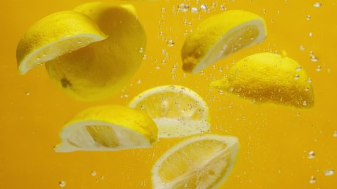 Close-up of falling sliced lemons into the water on orange background, making a cocktail of citrus fruits, drinking cold lemonade, shooting of carbonated water with sliced fruits. 