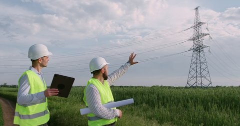 Two engineers look at an electronic tablet in front of a high-voltage power line, discuss transportation of electricity, and point their hand at electric poles in the open air.