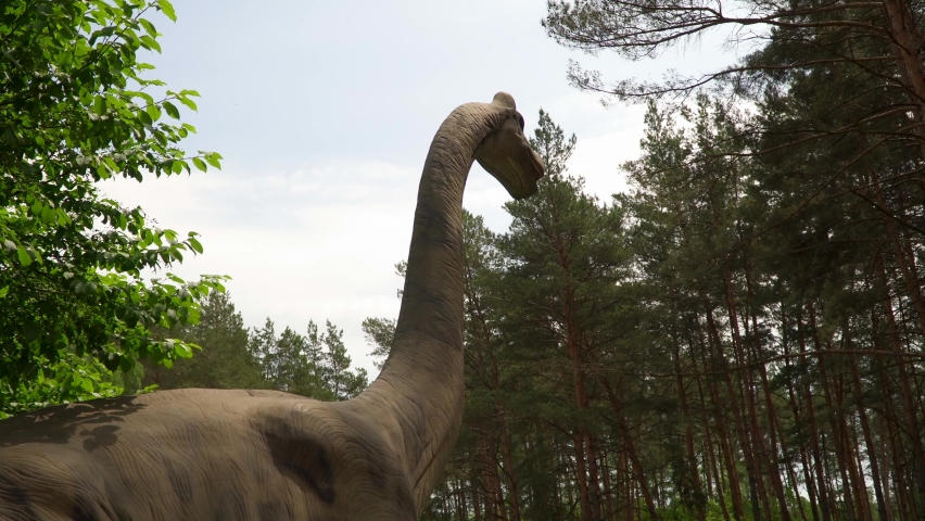 huge live dinosaur stands among trees in park. Dinosaur with crest in a green pine forest. Robotic model of dinosaur on display among trees. dinosaur chews and breathes. Large lizard of Mazazoi era.