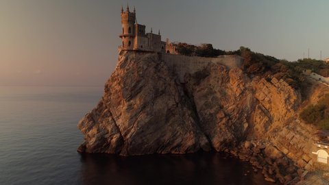 Aerial forward Swallow's Nest symbol of Crimea is an ancient castle palace on edge of cliff over Black sea at scenic orange sunset. Architectural monument Russia Ukraine Yalta Alupka. Travel landmark