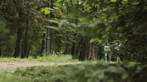 Wide shot of two senior athletic female joggers wearing sport clothes and sneakers having a run and talking in woods
