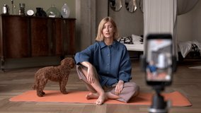Woman speaking in front of the phone, puppy walking around, yoga practicing sitting on mat in living room. Concept of online courses and sports blogger recording a video