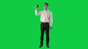 Business man with face mask streaming video on cell phone waving hand. Full length on green screen chroma key background