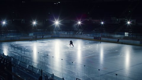 Ice Hockey Rink Arena: Professional Player Training Alone. Skates, Dribbles with Stick, Shooting, Hitting the Puck. Determined Athlete with Desire to Win, Be Champion. Cinematic Static Long Shot