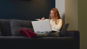 Ginger student with freckles is using a laptop doing her homework on a couch at home