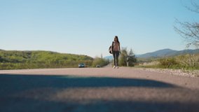 A young pretty woman with a backpack, with a tattoo on her hands, walks along the road. Lower angle. The concept of local hitchhiking.