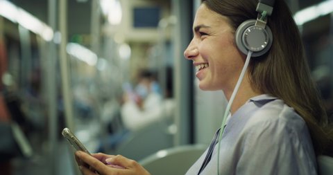 Cinematic shot of young smiling woman listening to the music with smartphone while traveling by train in subway. Concept of transportation, technology, connection, communication, lifestyle.