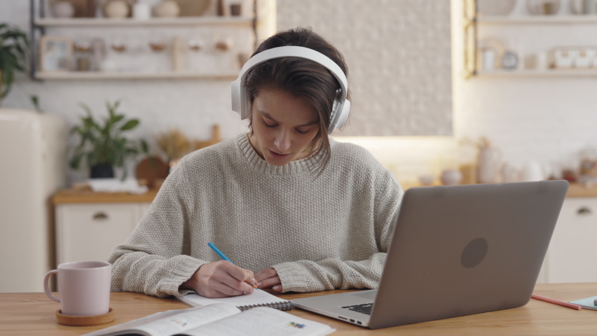 Pretty young woman in wireless headphones writing in copybook while using modern laptop at home. Female student spending free time at home for distance learning. Royalty-Free Stock Footage #1073896553