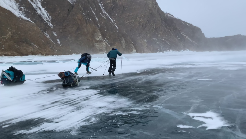 Two men are blown away by a hurricane wind with a blizzard during a hike across frozen Lake Baikal with sledges equipped with backpacks in winter. Storm on Lake Baikal in winter | Shutterstock HD Video #1073913686