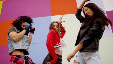 Three young beautiful and happy girls dancing , moving , looking at camera . Stylish bloggers in sport outfit  having fun , jumping and shaking body in front of colorful background wall in slow motion