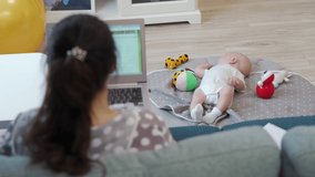 Back view of mother uses laptop while baby lies on activity play mat at home, lady working on laptop online, 3 month old baby boy playing with toys. High quality 4k footage