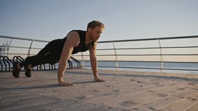 Sporty lifestyle concept. Zoom in and out shot of muscular man athlete practicing push ups outdoors, training on pier at seaside early at morning, slow motion