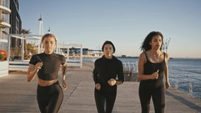 Three diverse women friends practicing morning jog workout, running on pier together early in morning at seaside, slow motion