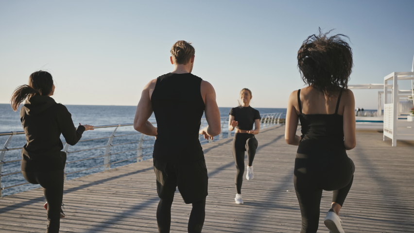 Intensive fitness training. Group of active diverse people practicing outdoor cardio training with coach, running on place on pier at seaside, tracking shot