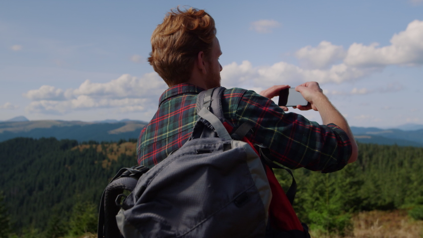 Focused guy with smartphone standing on top of mountain. Back view ginger man making panoramic photo of landscape on mobile phone. Male hipster taking video on cellphone during summer vacation