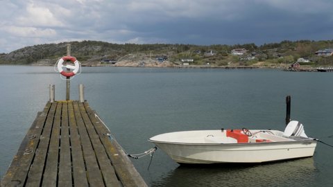 A lifebuoy is attached to the end of a jetty. A smaller boat with an outboard motor is moored at the jetty.  Dark clouds can be seen in the sky. The sea, rocks and houses can be seen in the background