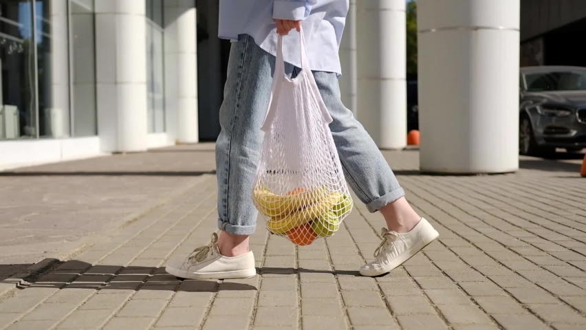 Woman in jeans walking and holding white mesh bag with fruits. Eco friendly, reusable shopping bag. Oranges, apples, bananas in cotton knitted string bag. Zero waste and plastic free concept. | Shutterstock HD Video #1073942930
