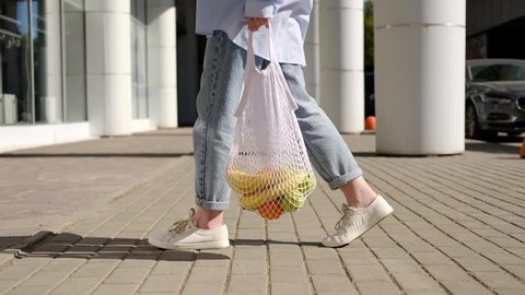 Woman in jeans walking and holding white mesh bag with fruits. Eco friendly, reusable shopping bag. Oranges, apples, bananas in cotton knitted string bag. Zero waste and plastic free concept.
