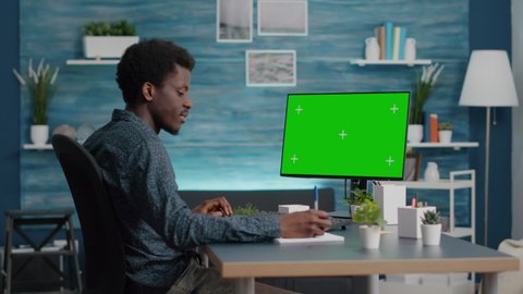 Black man typing and using personal computer with mock up chroma key display, isolated green screen PC for easy replacement. African american guy at home in bright living room