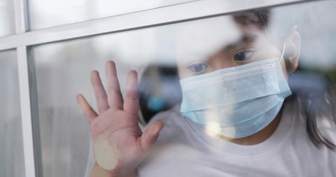 Slow motion scene, Through glass to little Asian girl in face mask looking out and place hand on window with Sadness during quarantine due to COVID alone at home.