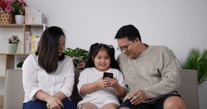 Asian family playing game on smartphone together in the living room. Happy mom and dad sit at home on couch with their little daughter on smartphone.