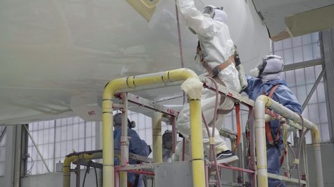 Indianapolis , IN , United States - 03 12 2021: Indianapolis, IN - March 12th, 2021: Two hispanic men in the aviation industry spray a top coat of gloss paint on the side of a commercial airplane in a