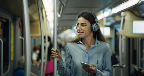 Cinematic shot of young smiling woman listening to the music with smartphone while traveling by train in subway. Concept of transportation, technology, connection, communication, lifestyle.