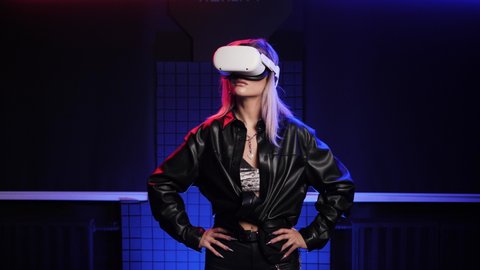Focused girl in VR glasses stands with hands on hips. Virtual reality helmets goggles, gamer person in AR headsets. Gaming people focus. Neon red and orange light color. Pretty young adult portraits.