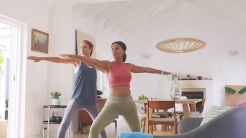 Two mature women doing warrior pose at home with stretched arms. Healthy mature woman doing yoga exercise with mixed race friend at home. Middle aged lady in sportswear exercising together.