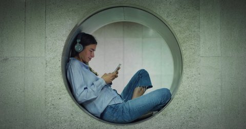 Cinematic shot of woman listening to music with headphones and using smartphone at subway station with passing people. Concept of urban, social media, technology, connection, communication, lifestyle.