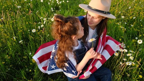 Cute young mother in hat and sweet small daughter holds usa United States flag sit on green meadow at summer day. Smiling free proud independent patriotic family feeling freedom, independence concept