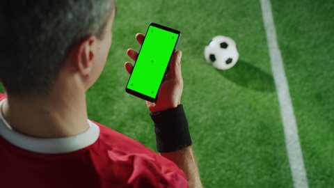 Professional Soccer Player Holding Smartphone with Green Screen Chroma Key with Tracking Markers. Elevated Top Down Over Shoulder Shot of Football Star Using Internet While Standing on Field